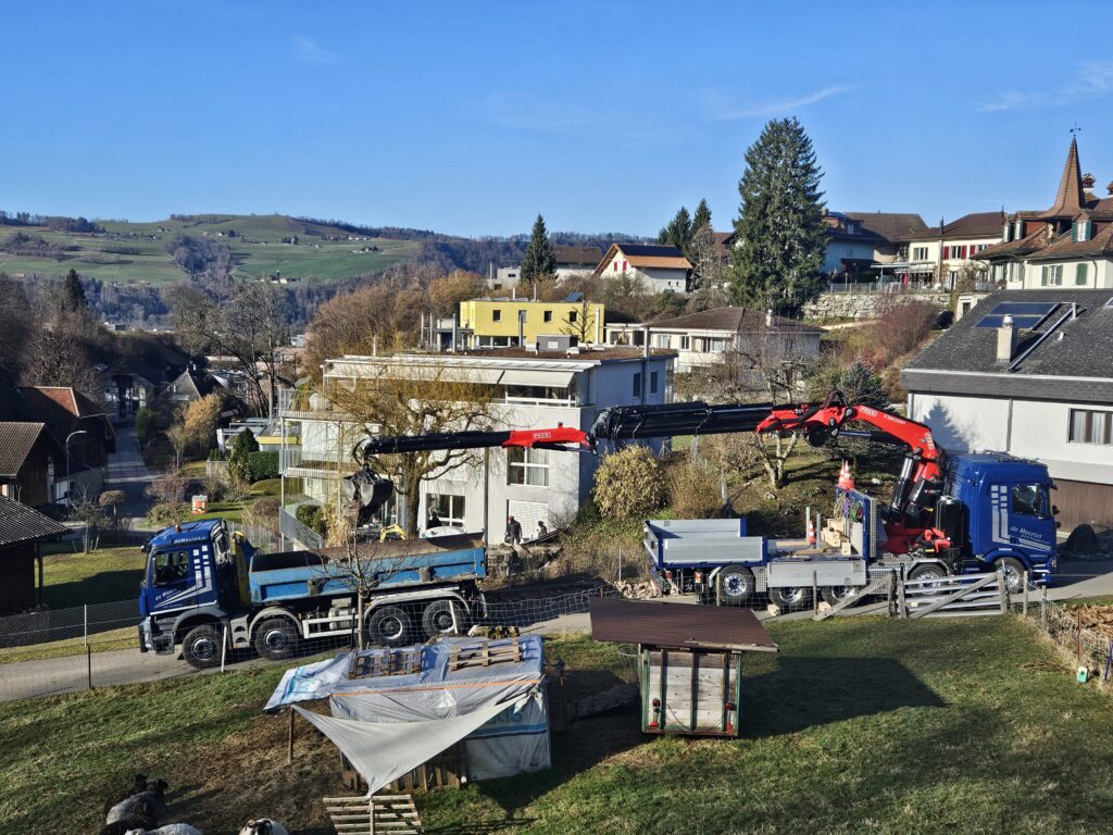 Zwei blaue Lastwagen mit ausgefahrenen roten Kranarmen laden Material vor Wohnhäusern in einer sonnigen Hügelsiedlung.