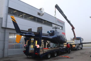 Dunkelblauer Helikopter wird mit Kran auf Tieflader vor Gebäude mit Schriftzug remund carrosserie verladen; Arbeiter dabei.