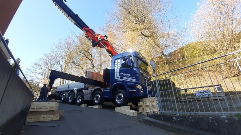 Blauer Lkw der Firma de Meuron mit ausgefahrenem roten Kranarm, auf Holzunterlagen stabilisiert an einer steilen Straße.