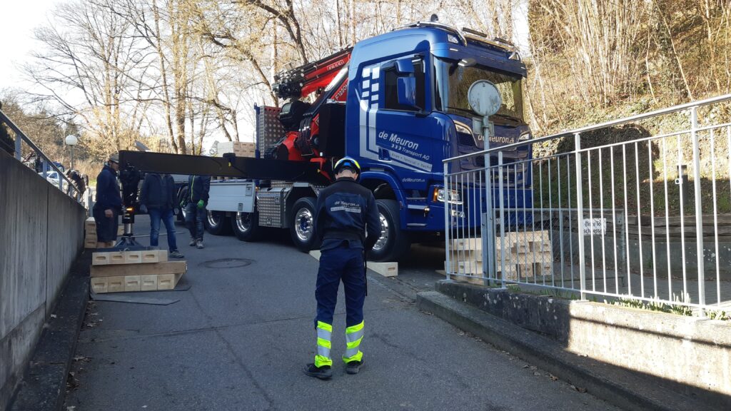 Mann in Warnhose vor blauem de Meuron-Lkw mit ausgefahrenem Kran, Arbeiter und Holzauflagen an schmaler Straße