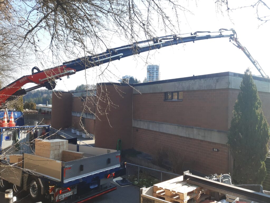 Ausgefahrener Kranarm über einem flachen Backsteingebäude, Lkw mit Holzkisten und Baustellenmaterial im Vordergrund.
